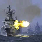 A ship at sea firing artillery while three other ships follow behind.