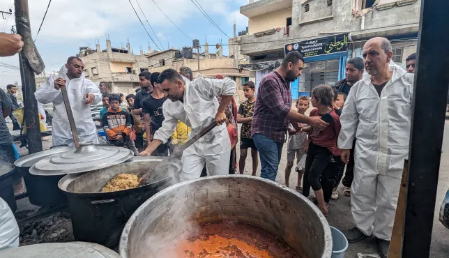 Workers in white overalls stir large vats of food in the community kitchen, people and buildings in the background