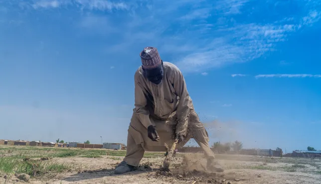 A man works the dry, dusty soil with a hoe under a bright blue sky in a rural area, with modest shelters visible in the background — a scene reflecting resilience in the face of harsh living conditions.