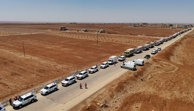 A long humanitarian aid convoy stretches along a rural road in Sweida, Syria, in July 2025. The convoy includes dozens of white vehicles marked with the Red Cross and Red Crescent emblems, along with large trucks carrying relief supplies. The road runs through a dry, reddish-brown landscape with sparse vegetation and scattered buildings in the distance. Aid workers in red uniforms walk near the vehicles under a clear blue sky.