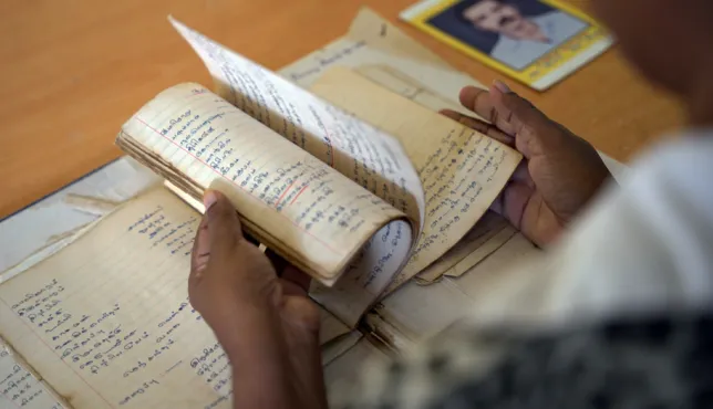 A person flips through the pages of an old, handwritten notebook. Several other aged notebooks lie on the table, along with a photograph of a man placed in the background.