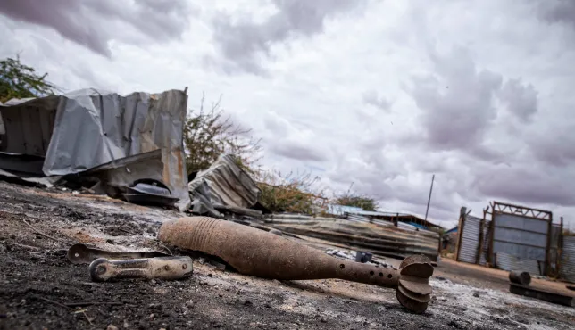 A rusted, unexploded mortar round lies on scorched ground in an abandoned area of Gedo’s Luuq district, Somalia. Behind it, twisted metal sheets and damaged structures sit under a heavy, overcast sky, highlighting the destruction left by recent fighting.