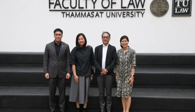 Four people standing and posing for a group photo in front of a wall that reads “Faculty of Law, Thammasat University,” with the university’s emblems displayed above them.
