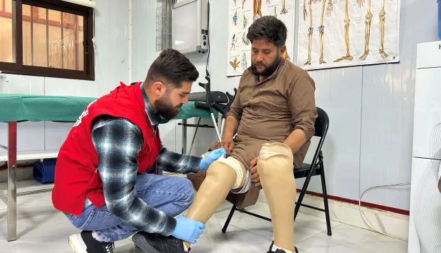 Joumaa, seated on a chair, wears two lower-limb prostheses as an ICRC staff member in a red vest kneels to adjust the fit. They are in a clinical rehabilitation room with anatomical posters on the wall.