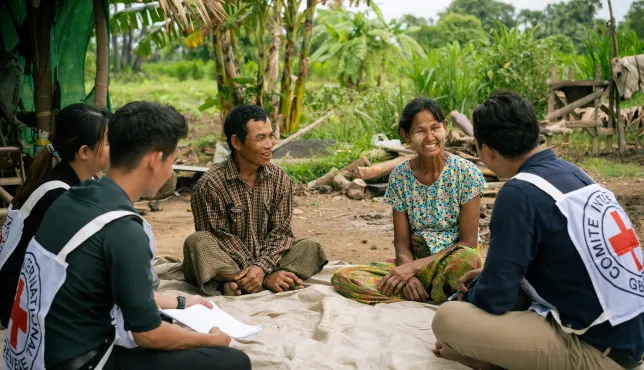 ICRC staff and volunteers sit with two local community members on a mat outdoors in a rural setting