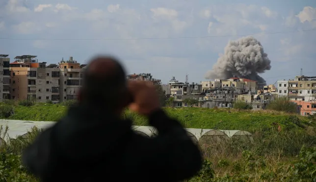 A person stands in the foreground, blurred and seen from behind, holding a phone while looking toward a city where a large plume of smoke rises above buildings in the distance.
