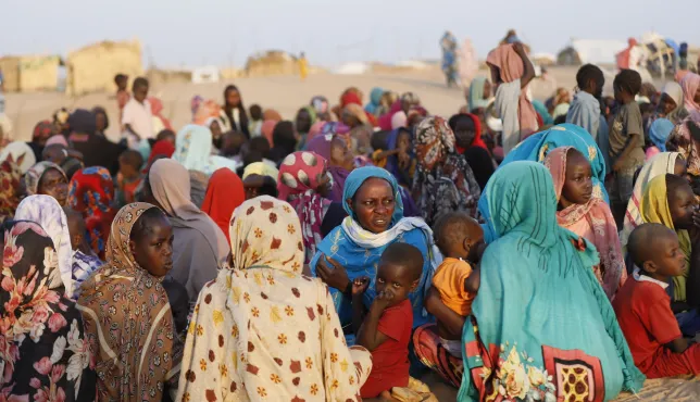Crowd of women and children gathered outdoors in Tawila, North Darfur, many seated on the ground wearing colorful headscarves and traditional clothing, with a few people standing in the background against a dry, sandy landscape.