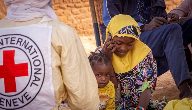A woman wearing a yellow headscarf sits and speaks on a phone, with a young child beside an ICRC vest in Anderamboukane, Mali, as people gather in the background outdoors.