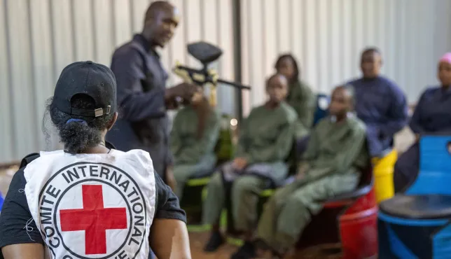 An ICRC staff member oversees a practical session of Law Students during an armed conflict simulation excersise on IHL. She is looking at an instructor holding a mock rifle as students are seated looking attentively looking at the instructor.