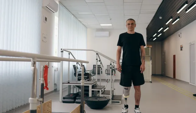 A man with two lower-leg prosthetic limbs stands in a rehabilitation centre beside parallel bars used for gait training. Exercise equipment is visible in the background of the bright therapy room.
