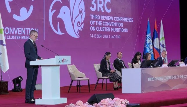 ICRC Vice-President Jürg Lauber speaks at the Third Review Conference of the Convention on Cluster Munitions, with panelists seated on stage and conference branding and flags in the background.