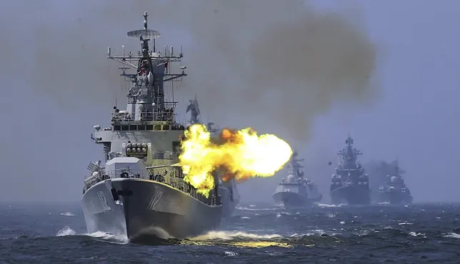 A ship at sea firing artillery while three other ships follow behind.